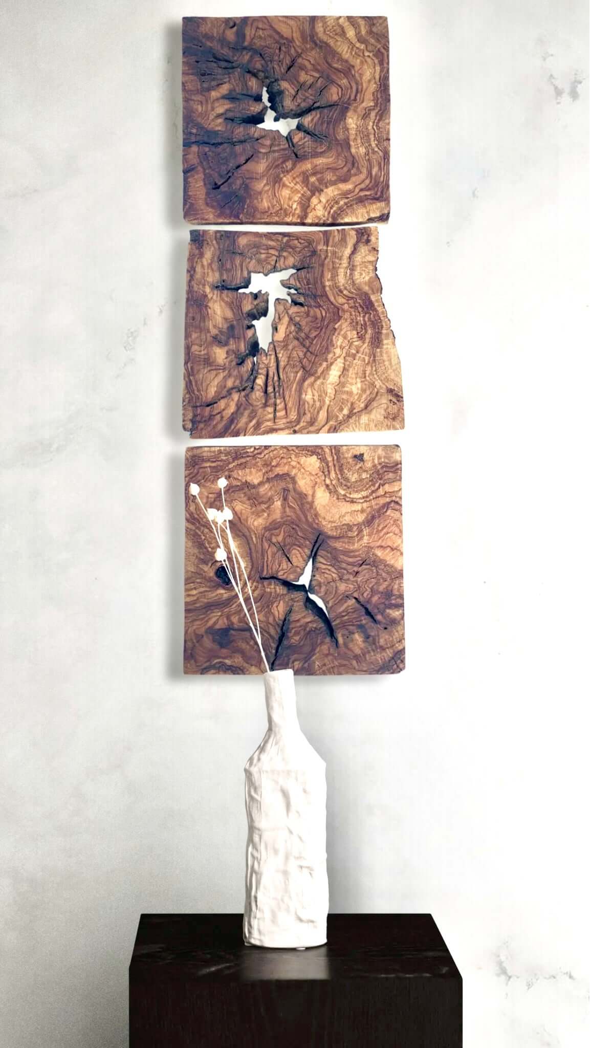 Three wooden panels with abstract designs on a white wall, with a white vase on a black pedestal.
