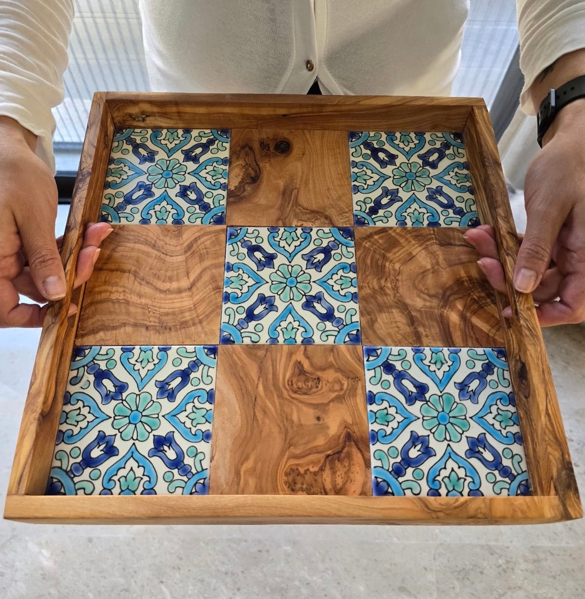 Wooden tray with a checkered pattern of blue and green tiles held by two hands.