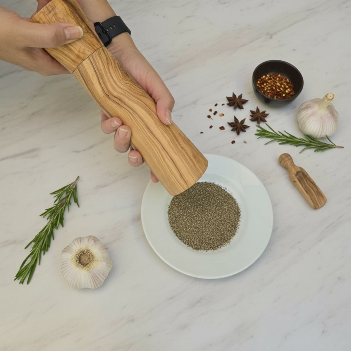 Hand using a wooden pepper grinder on a white plate with spices and herbs on a light wood surface.
